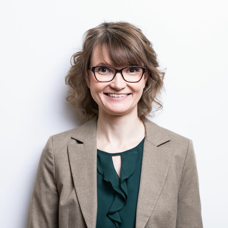 Portr&auml;t von Claudia Schafranski mit Brille, lockigem Haar und beigem Blazer vor hellem Hintergrund